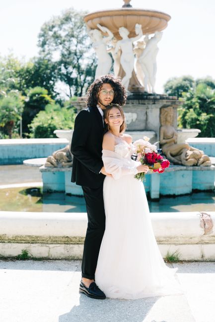 A floral gift consisting of a vibrant bouquet of deep pink and white roses, accented with greenery, held by a smiling bride in a white wedding dress. The bouquet features a mix of fresh, textured peta