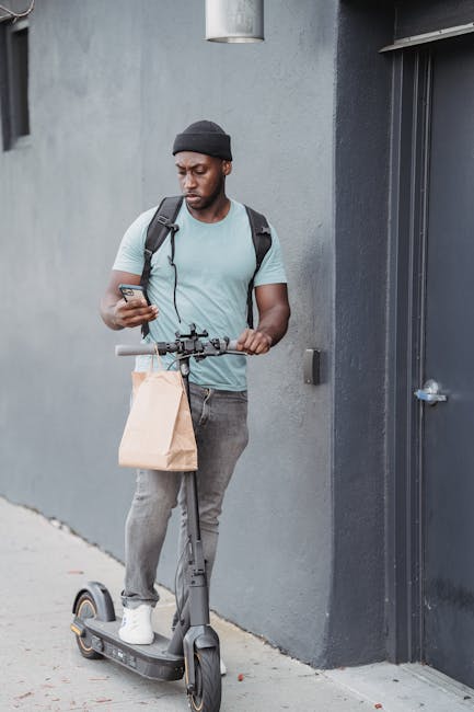 A young man with dark skin, wearing a black beanie, light grey t-shirt, and grey jeans, standing outside against a grey concrete wall near a dark grey door. He is holding a smartphone in one hand and 